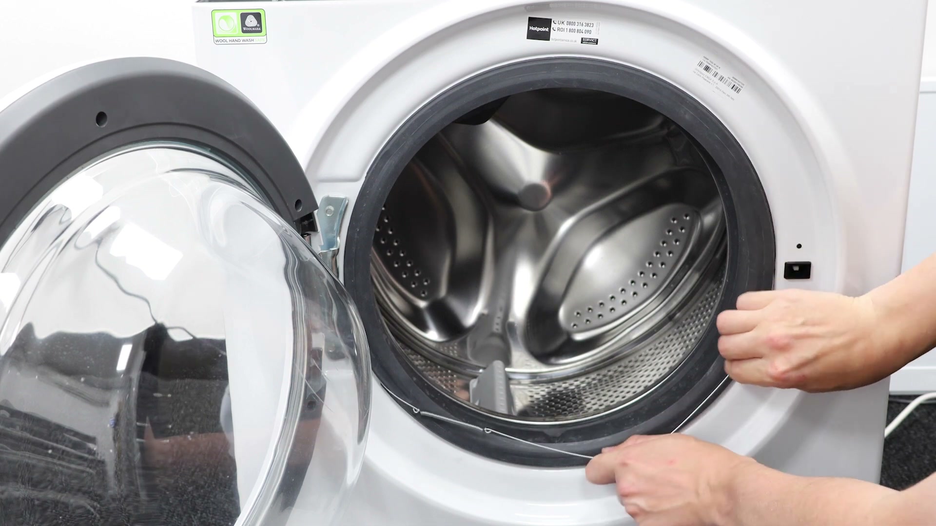 Hands refitting the outer clamping band ring spring around the front of a Hotpoint washing machine door seal